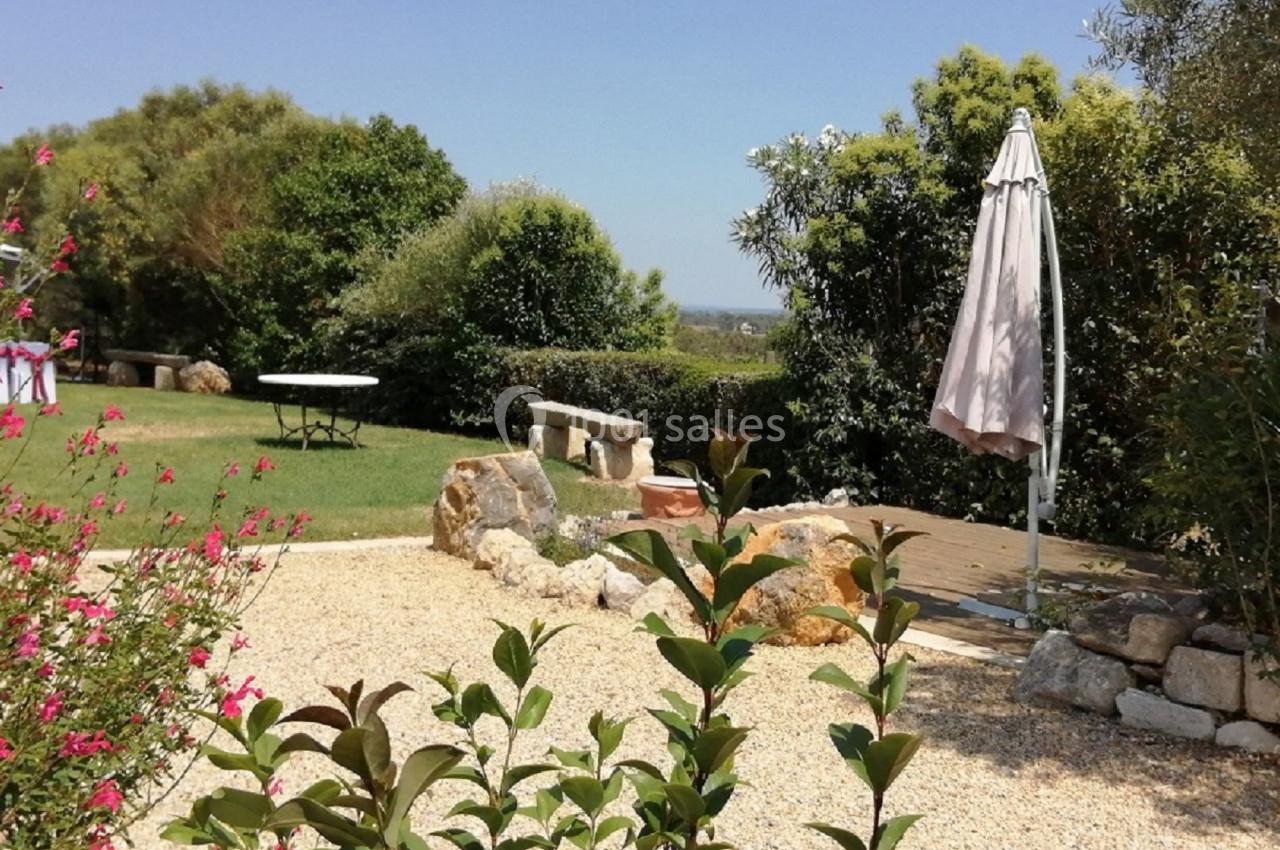 Jardin avec terrasse en gravier, parasol fermé, table en arrière-plan et végétation luxuriante sous un ciel dégagé.