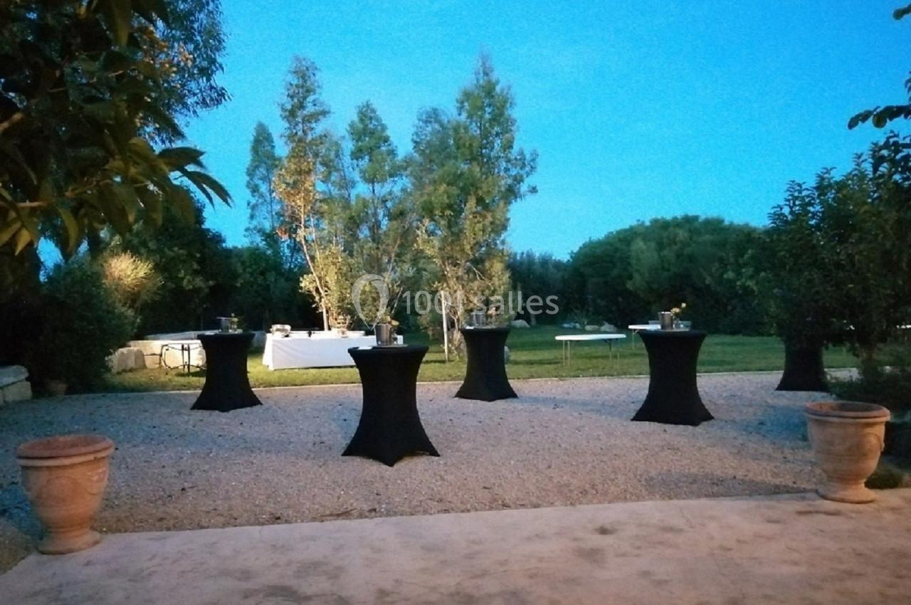 Espace extérieur aménagé avec des tables hautes noires sur une terrasse gravillonnée, entouré de verdure au crépuscule.