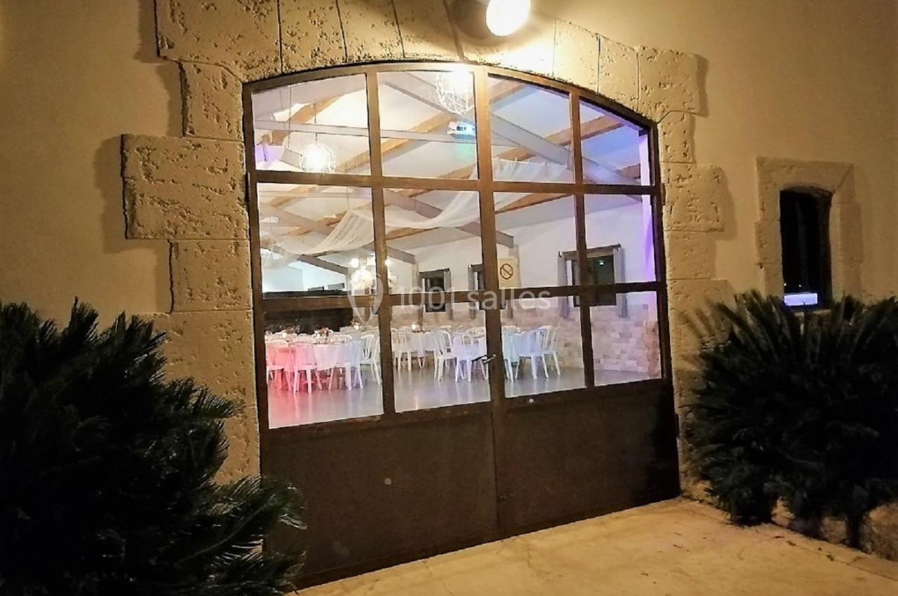 Grande porte vitrée en arc donnant sur une salle de réception éclairée, entourée de murs en pierre et de végétation.