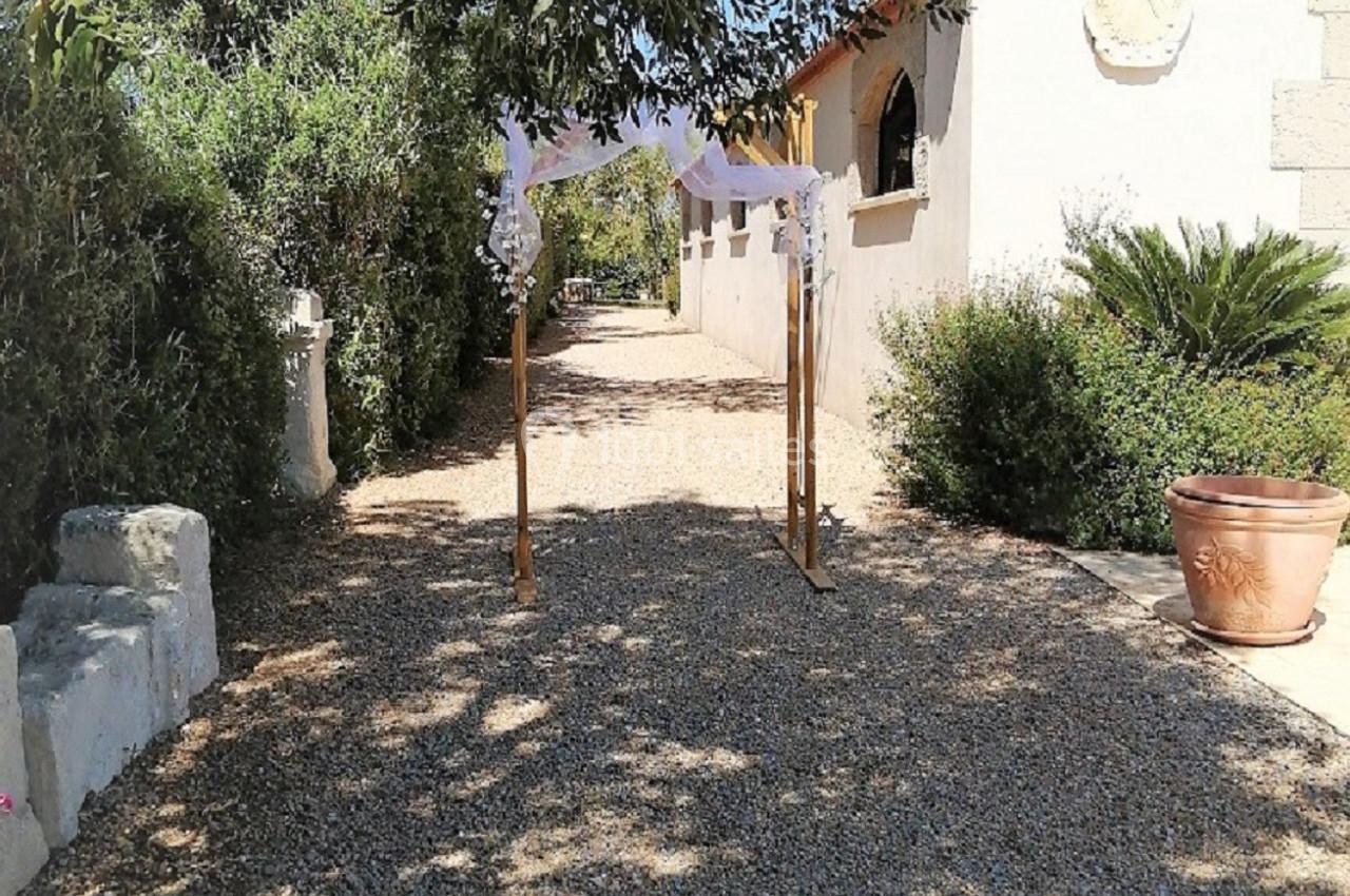 Allée en gravier bordée de végétation, avec une arche décorée de tissu blanc menant à un bâtiment clair.