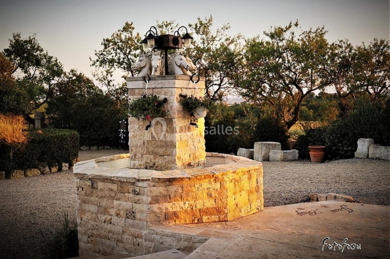 Fontaine en pierre rustique avec sculptures, entourée de végétation et éclairée par une lumière douce au coucher du soleil.