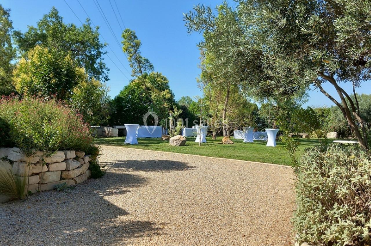 Allée en gravier menant à un jardin verdoyant avec des tables hautes blanches disposées sur une pelouse.