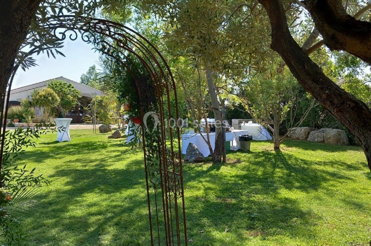 Vue d'un jardin verdoyant avec des tables dressées pour un événement, encadré par une arche métallique et des arbres.