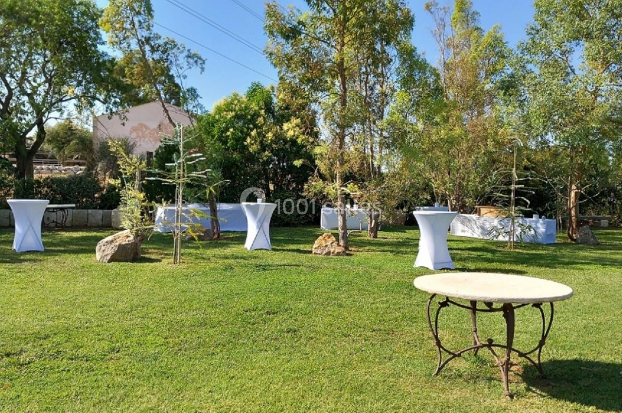 Jardin avec des tables hautes drapées de blanc, entouré d'arbres et de verdure sous un ciel dégagé.