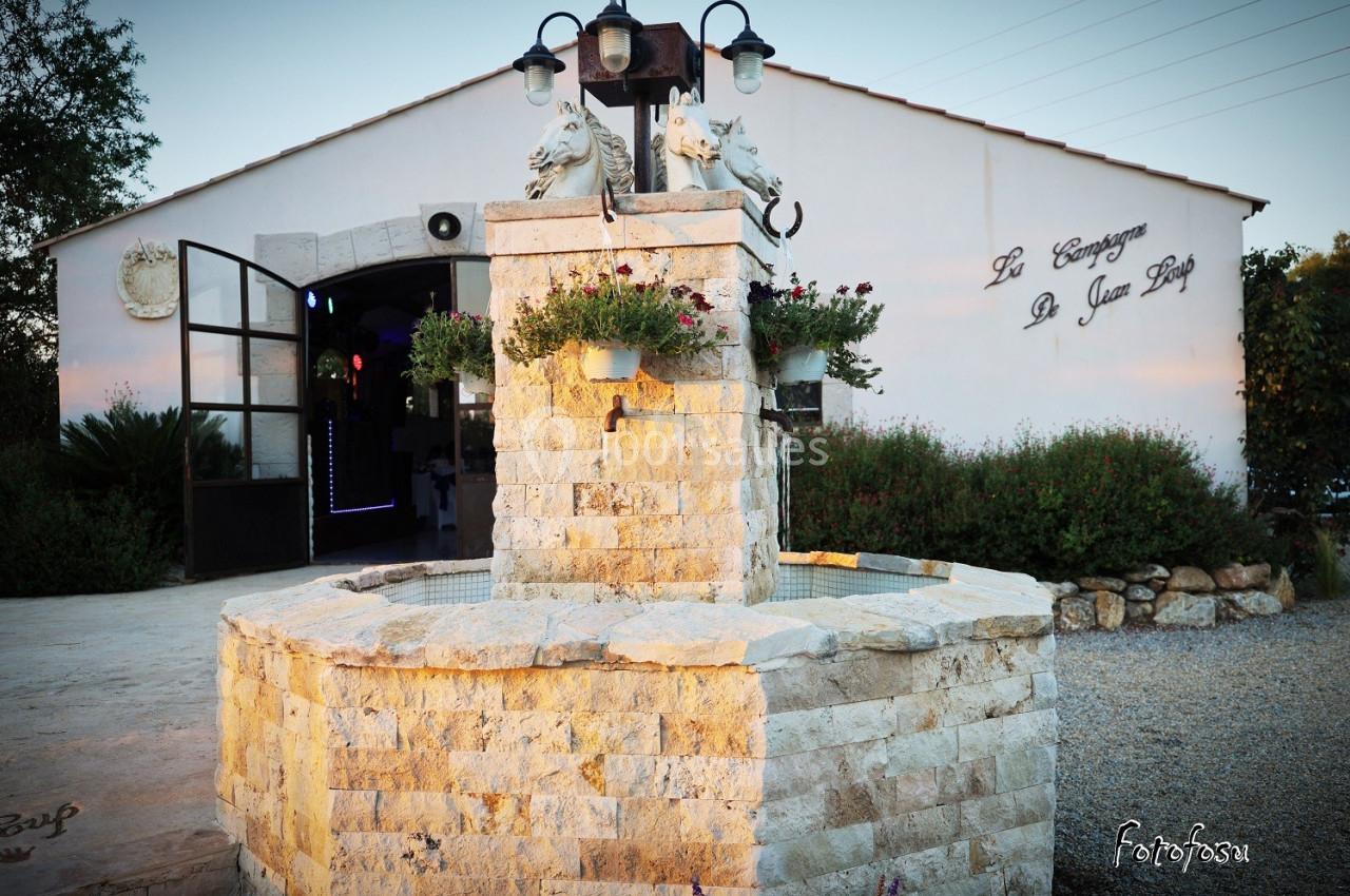 Fontaine en pierre ornée de sculptures de chevaux, devant un bâtiment blanc avec une entrée éclairée.
