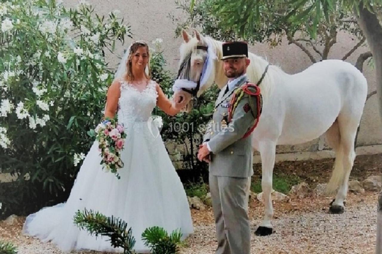 Un couple en tenue de mariage pose à côté d'un cheval blanc dans un jardin entouré de verdure.