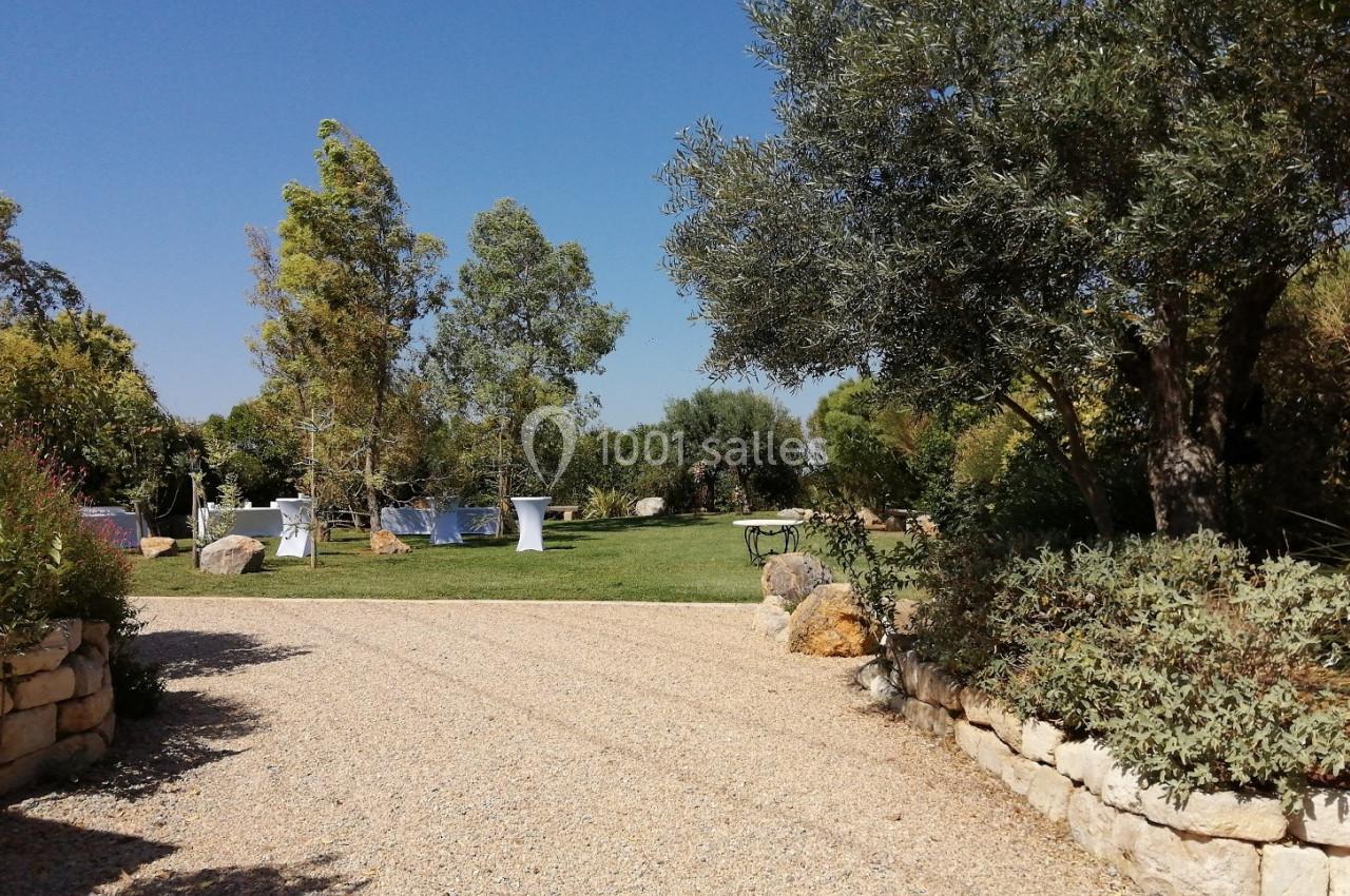Allée en gravier bordée de végétation menant à un jardin avec des tables dressées sous un ciel dégagé.