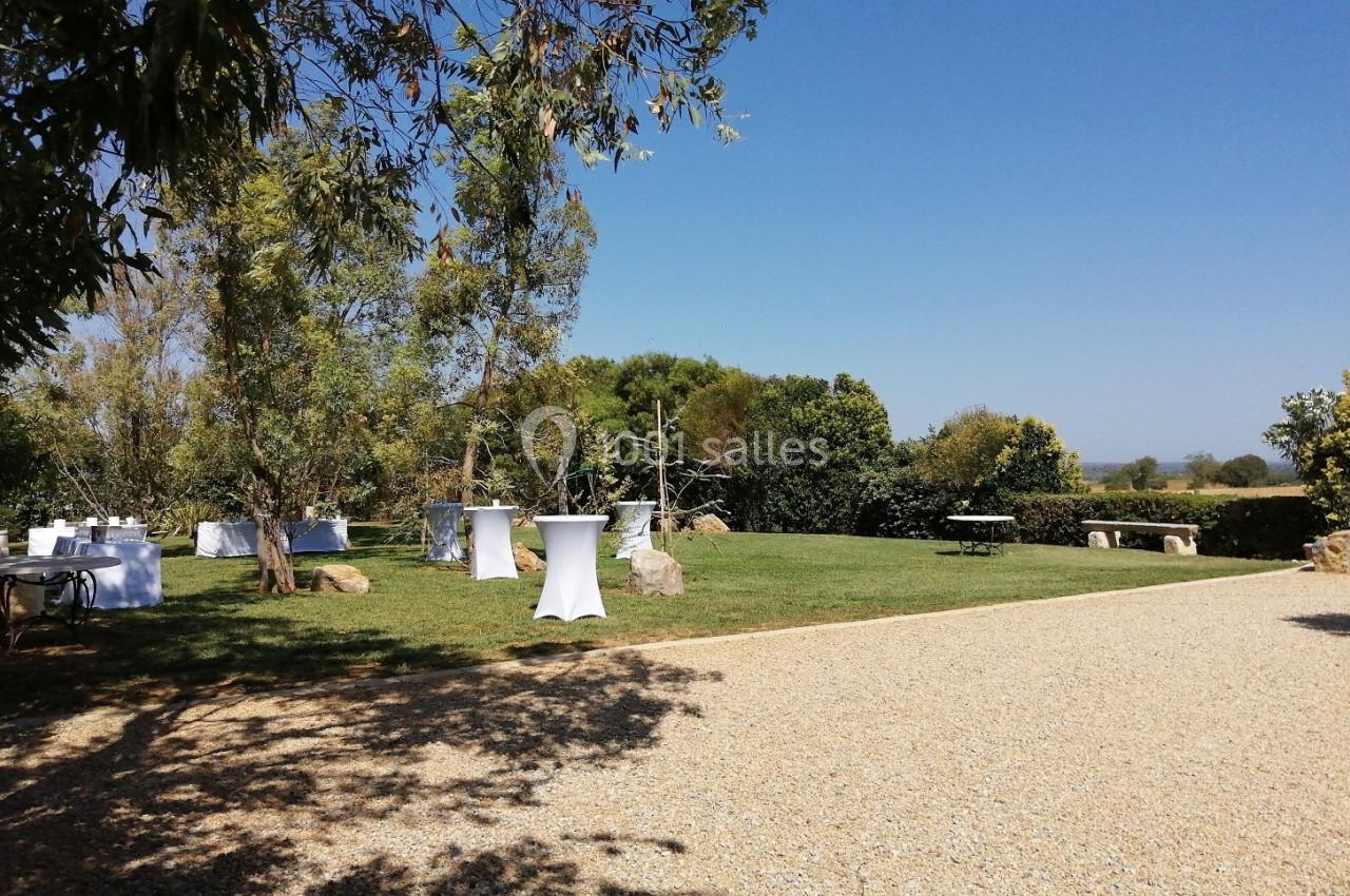 Espace extérieur avec pelouse, tables hautes blanches et allée gravillonnée, entouré de végétation sous un ciel bleu.