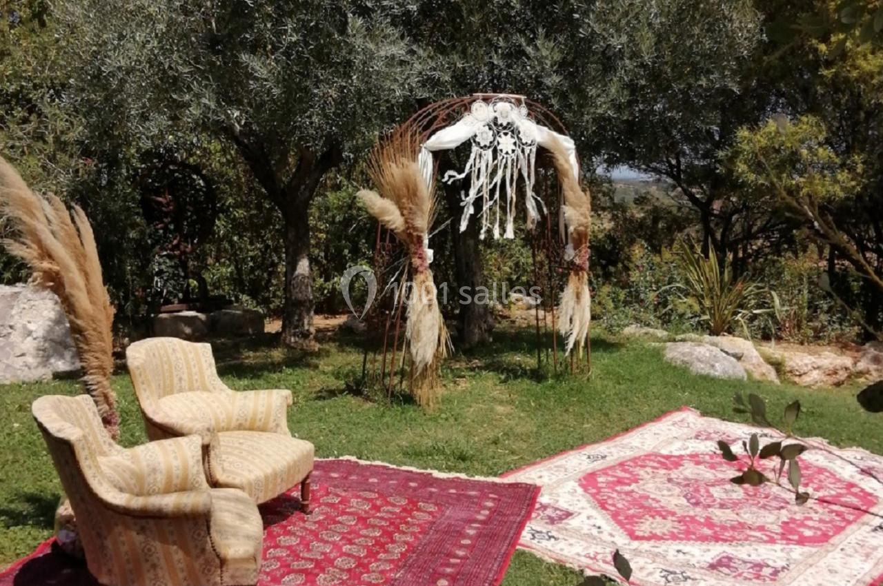 Deux fauteuils et des tapis rouges disposés sur une pelouse, sous une arche décorée de pampas et de macramé, près d'oliviers.