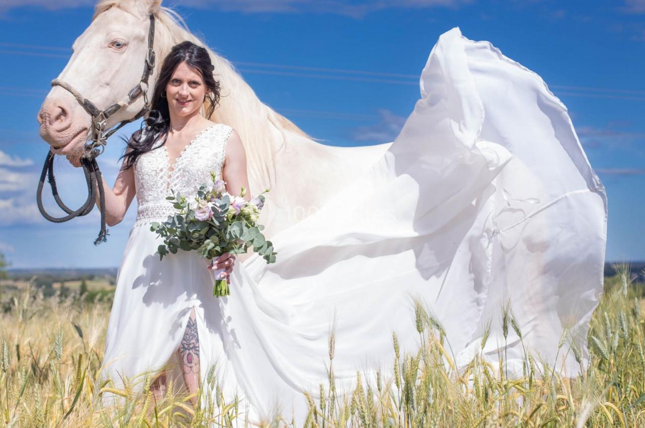 Femme en robe blanche tenant un bouquet, debout dans un champ avec un cheval clair sous un ciel bleu.
