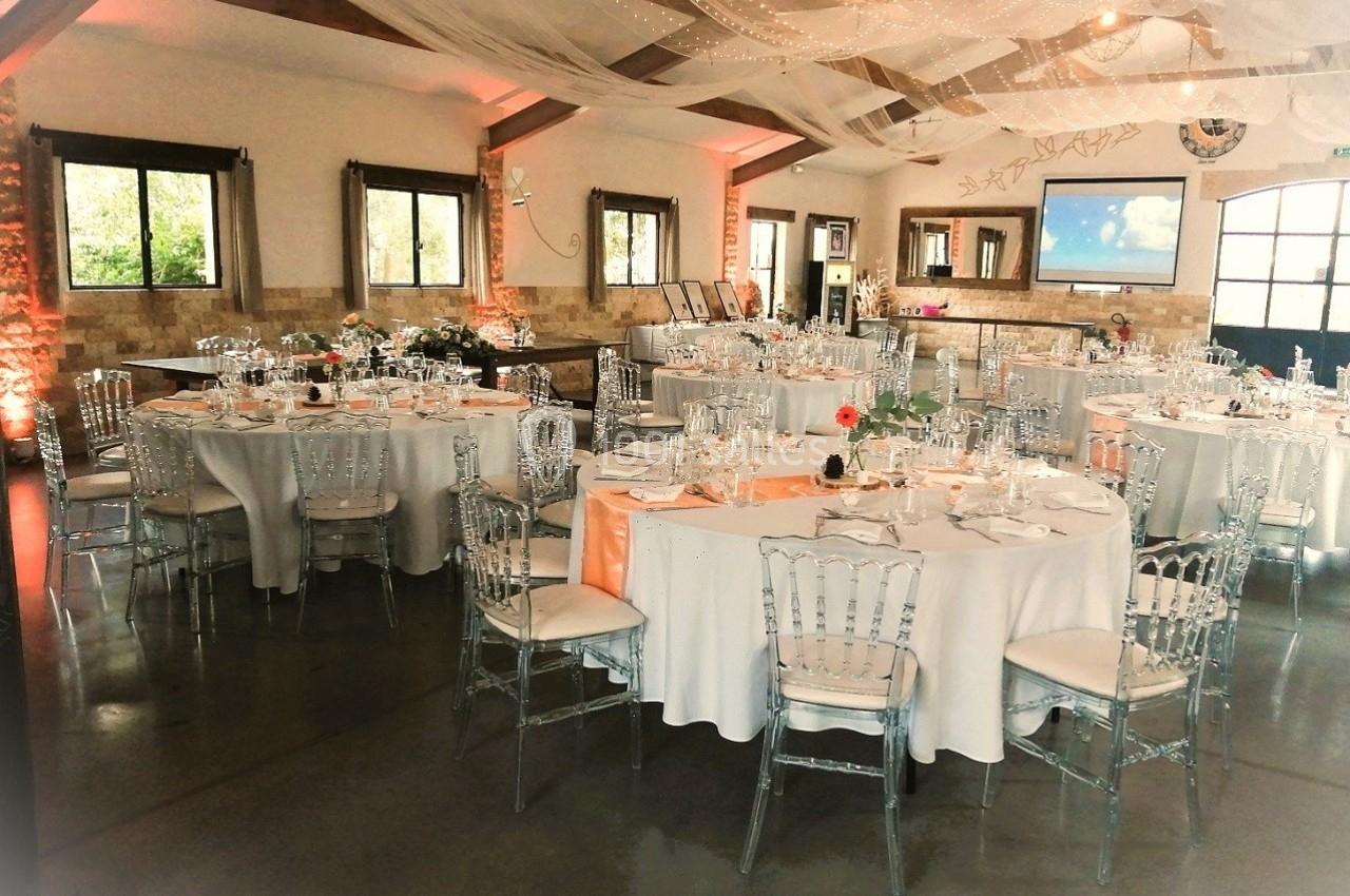 Salle de réception décorée avec des tables rondes dressées, des chaises transparentes et des voilages suspendus.