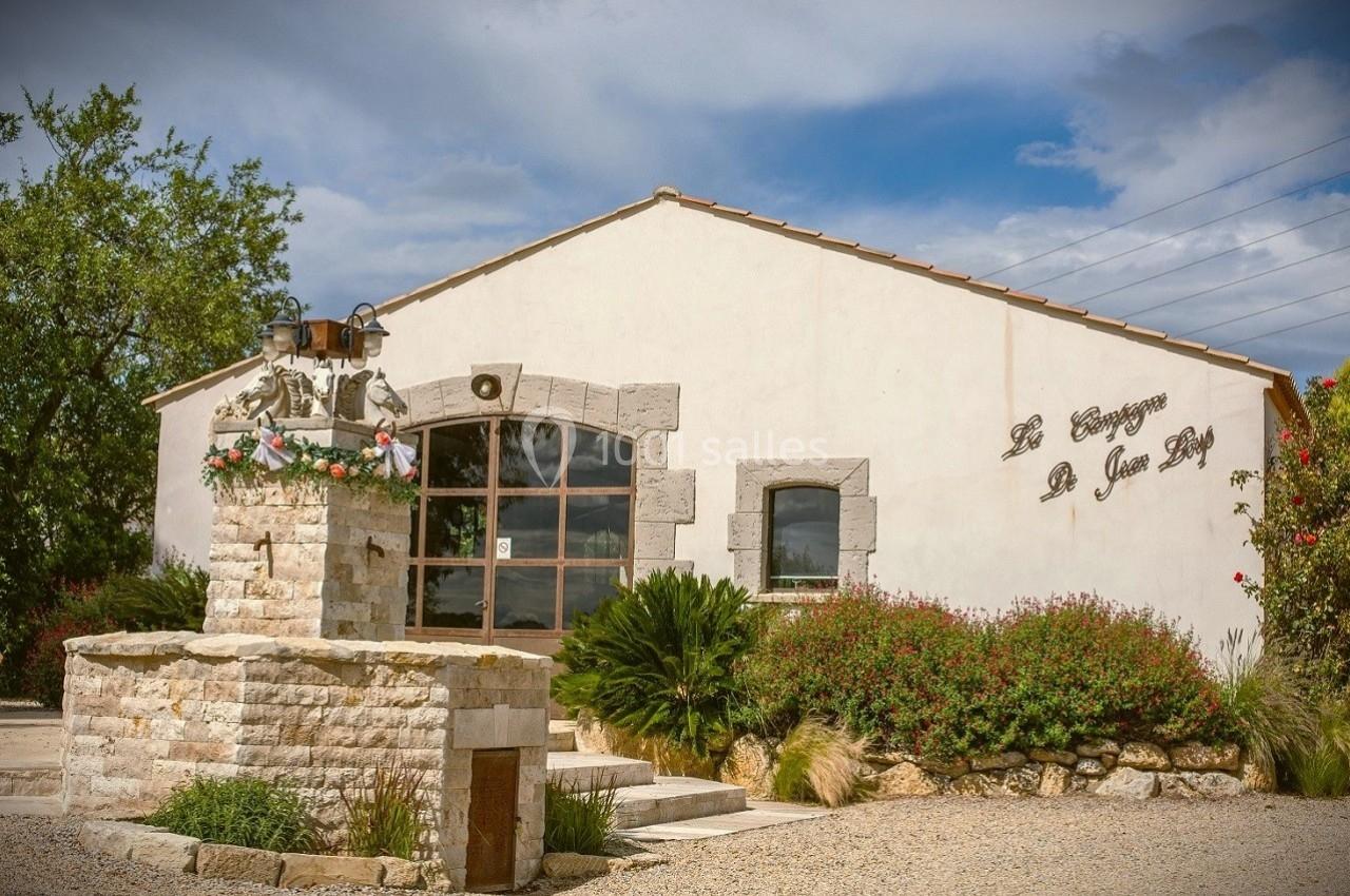 Bâtiment en pierre avec une fontaine fleurie au premier plan, entouré de végétation sous un ciel partiellement nuageux.