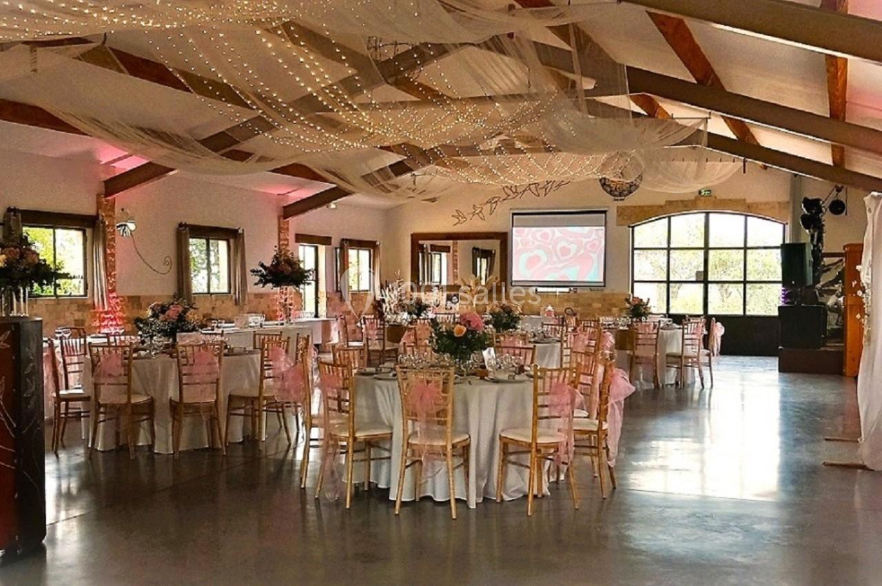 Salle de réception décorée pour un événement, avec tables rondes, chaises en bois, guirlandes lumineuses et voilages…