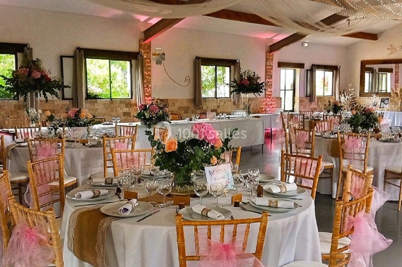Salle de réception décorée avec des tables rondes, nappes blanches, chaises en bois et arrangements floraux colorés.