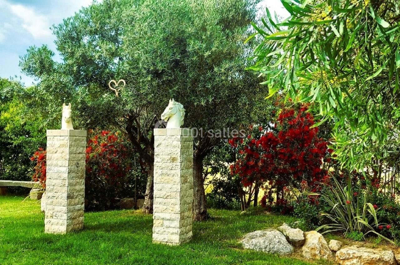 Deux statues de chevaux sur des colonnes en pierre dans un jardin verdoyant avec des arbres et des arbustes fleuris.