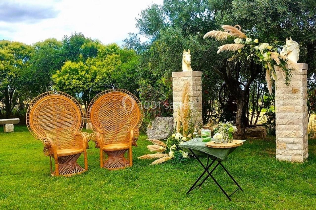 Deux fauteuils en rotin et une table décorée, installés dans un jardin verdoyant avec des colonnes ornées de fleurs.