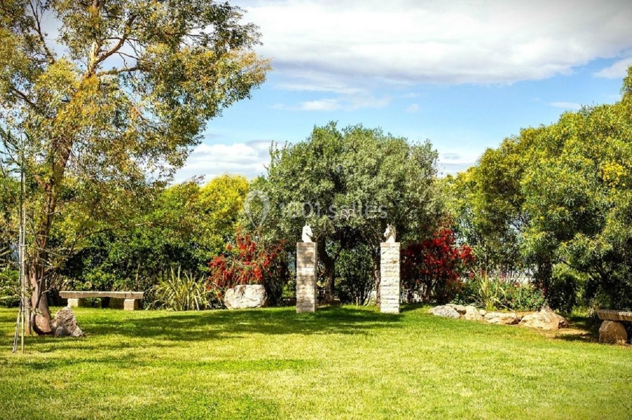 Pelouse verdoyante avec arbres, bancs en pierre et deux colonnes ornées de sculptures dans un jardin ensoleillé.