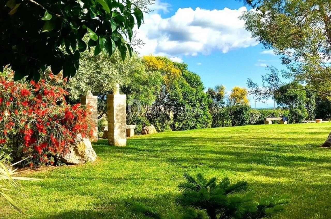 Jardin verdoyant avec pelouse, arbres, arbustes fleuris rouges et ciel bleu partiellement nuageux en arrière-plan.