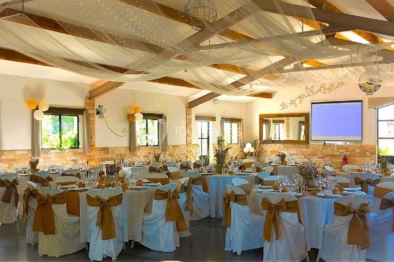 Salle de réception décorée avec des tables rondes, nappes blanches, chaises ornées de nœuds dorés et plafond drapé.