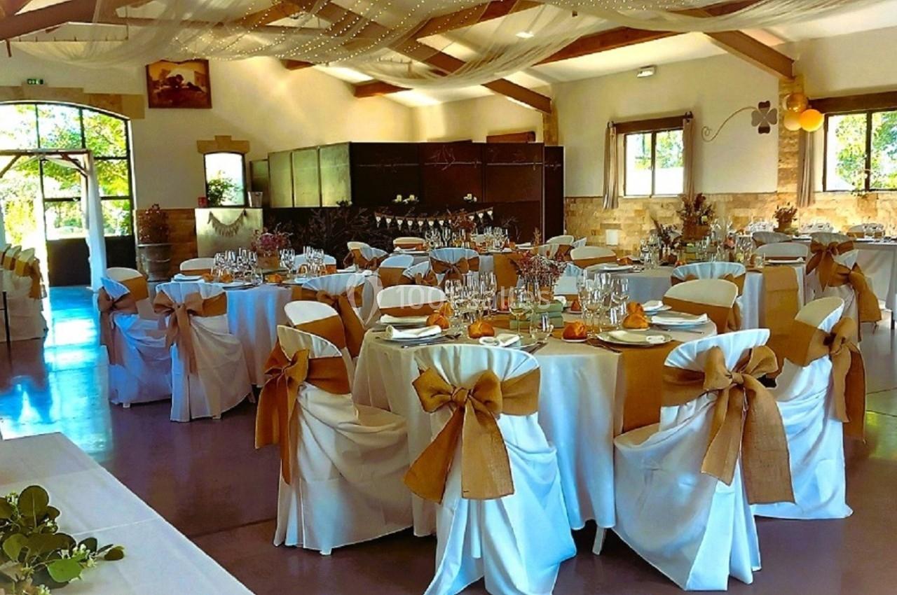 Salle de réception décorée pour un mariage, avec tables rondes dressées et chaises ornées de nœuds en tissu beige.
