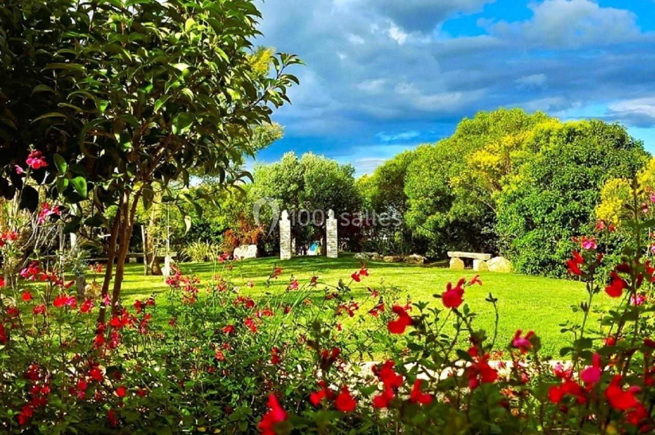 Jardin verdoyant avec fleurs rouges au premier plan, arbres et sculptures en pierre sous un ciel partiellement nuageux.