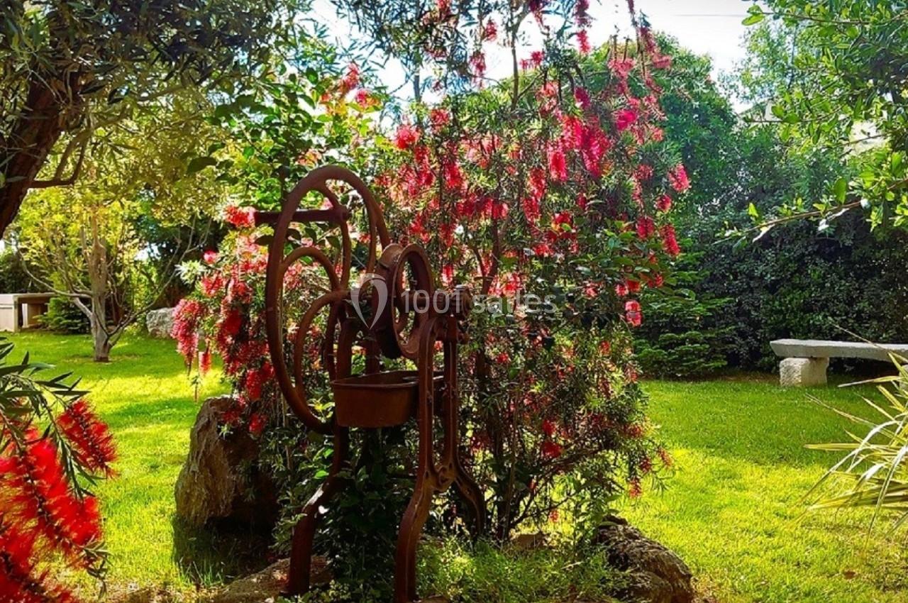 Ancienne pompe à eau en métal dans un jardin verdoyant avec des arbres et des fleurs rouges en arrière-plan.