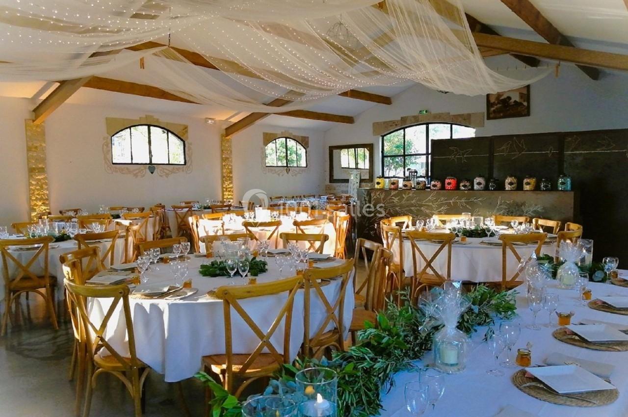 Salle de réception décorée pour un événement, avec tables rondes dressées, chaises en bois et voilages suspendus.