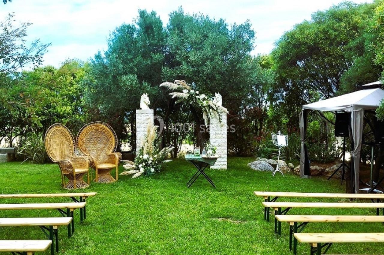Décoration de cérémonie en plein air avec arche fleurie, fauteuils en osier et bancs en bois sur une pelouse.