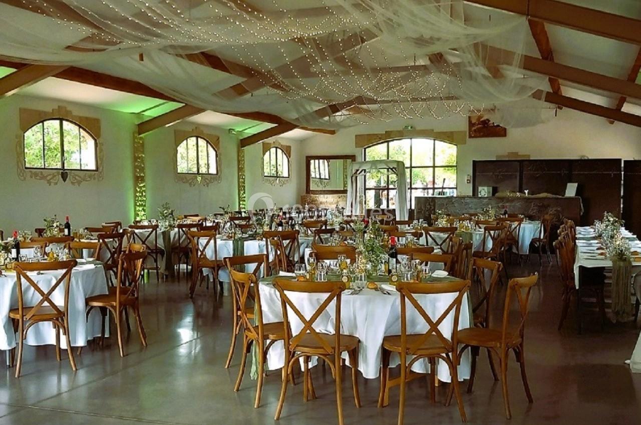 Salle de réception décorée avec des tables rondes dressées, des chaises en bois et un plafond orné de voilages et guirlandes…