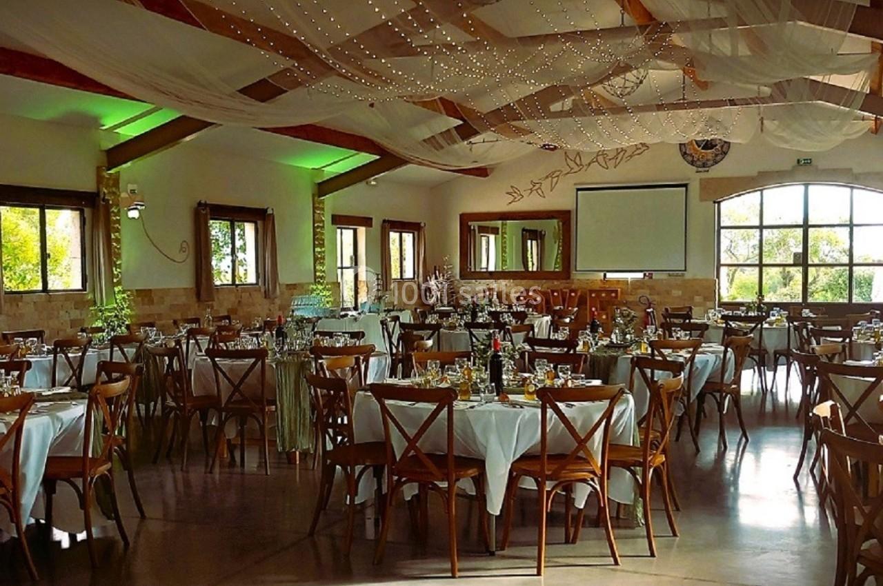 Salle de réception décorée avec des tables rondes dressées, des chaises en bois et des guirlandes lumineuses suspendues.