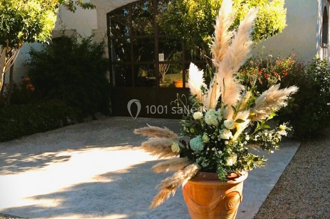Pot en terre cuite avec des fleurs et des herbes décoratives devant une bâtisse blanche entourée de végétation.
