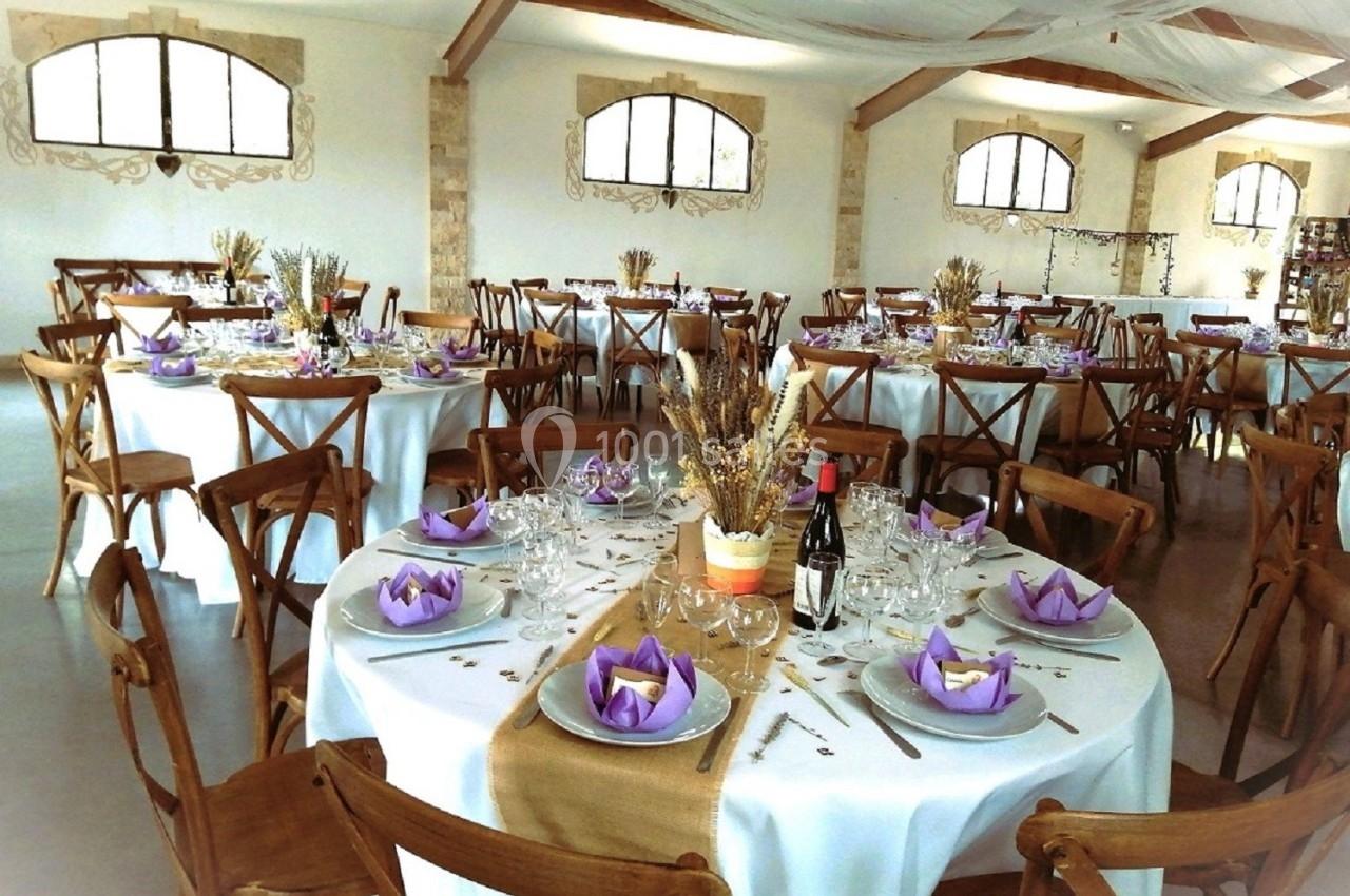 Salle de réception décorée avec des tables rondes, nappes blanches, serviettes violettes et centre de table floral.