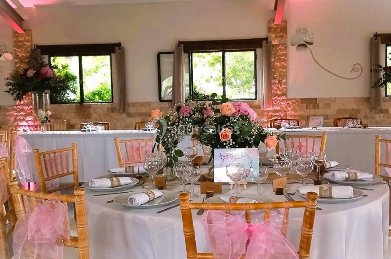 Salle de réception décorée avec des tables rondes, nappes blanches, chaises ornées de rubans roses et compositions florales.