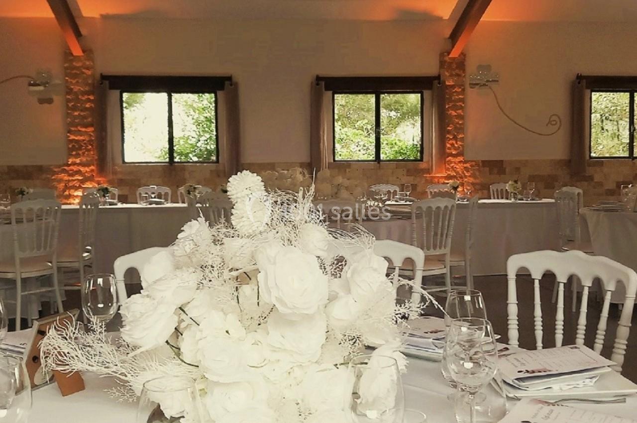 Salle de réception décorée avec des tables rondes, nappes blanches, bouquets de fleurs et éclairage chaleureux.