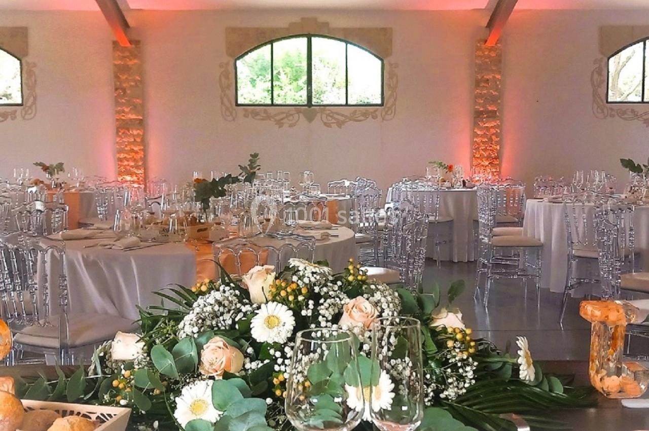 Salle de réception décorée avec des tables rondes, nappes blanches, chaises transparentes et compositions florales.