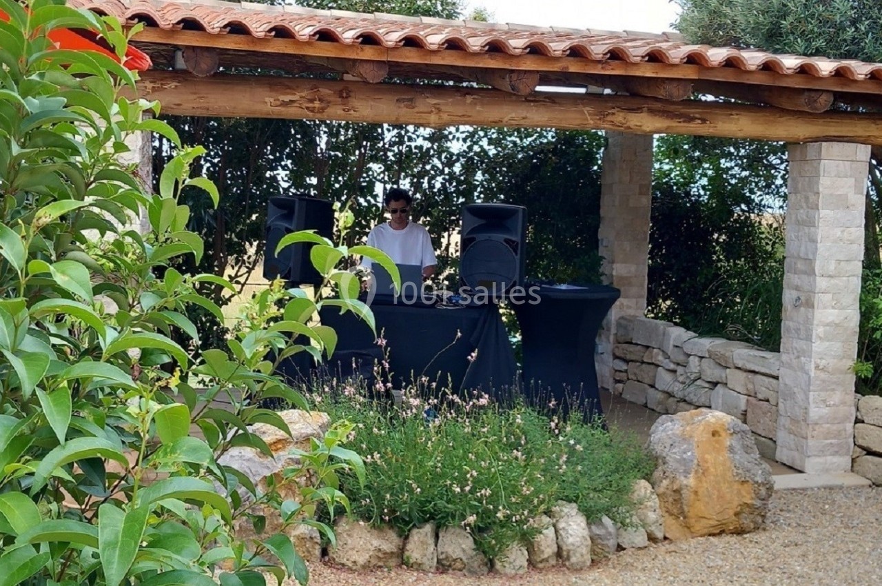 Un DJ mixe sous une pergola en pierre entourée de végétation dans un jardin méditerranéen.