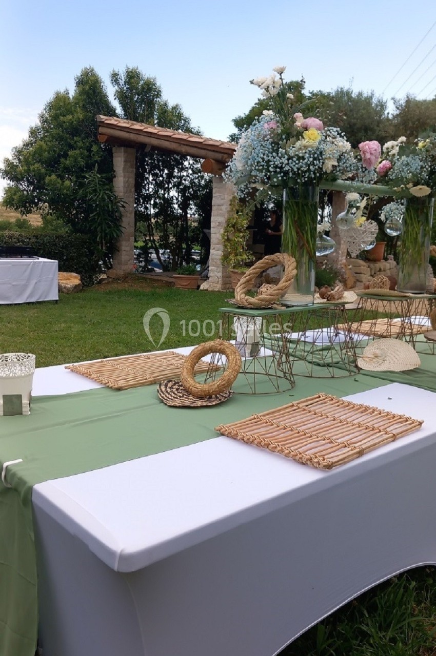 Table décorée en extérieur avec nappes blanches, chemins de table verts, fleurs et éléments en osier dans un jardin.