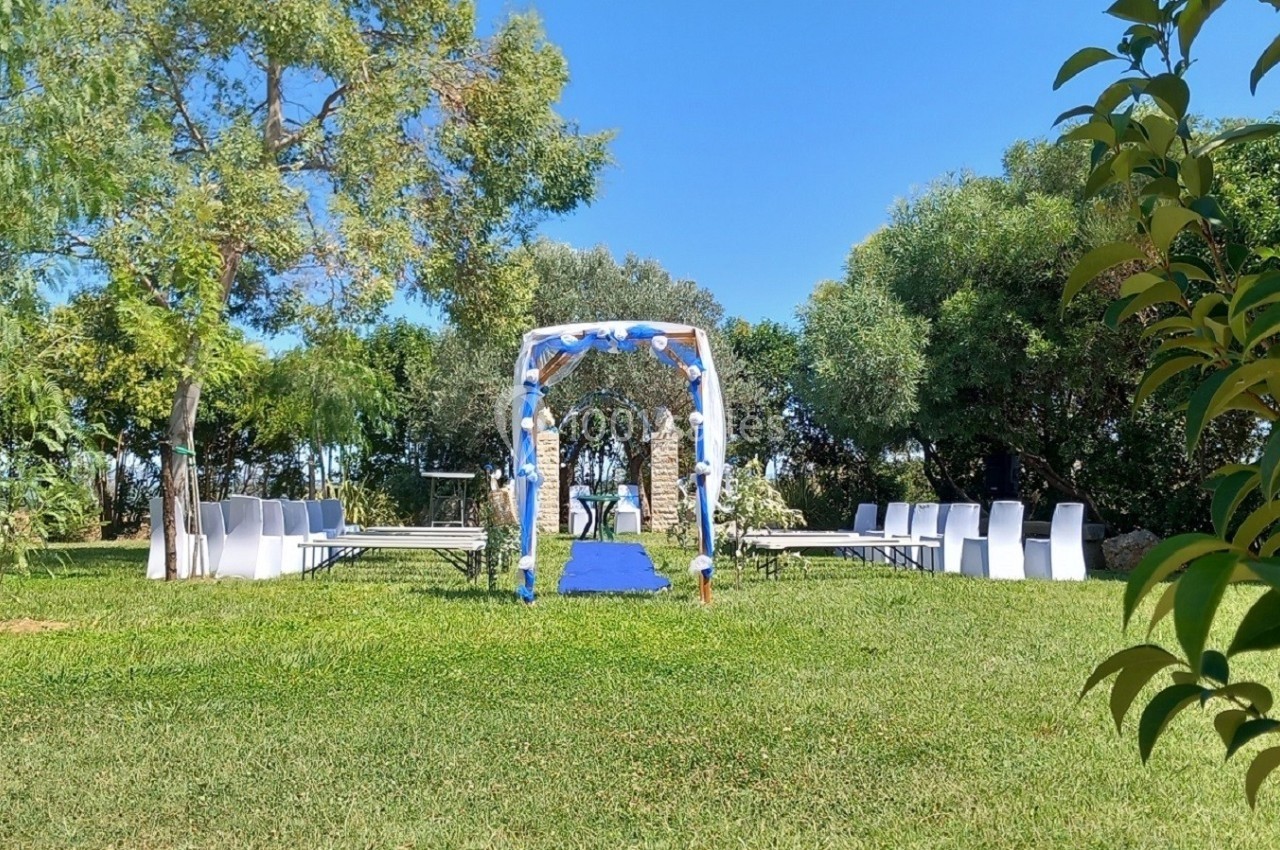 Allée centrale avec tapis bleu menant à une arche décorée pour une cérémonie en plein air, entourée de chaises blanches.