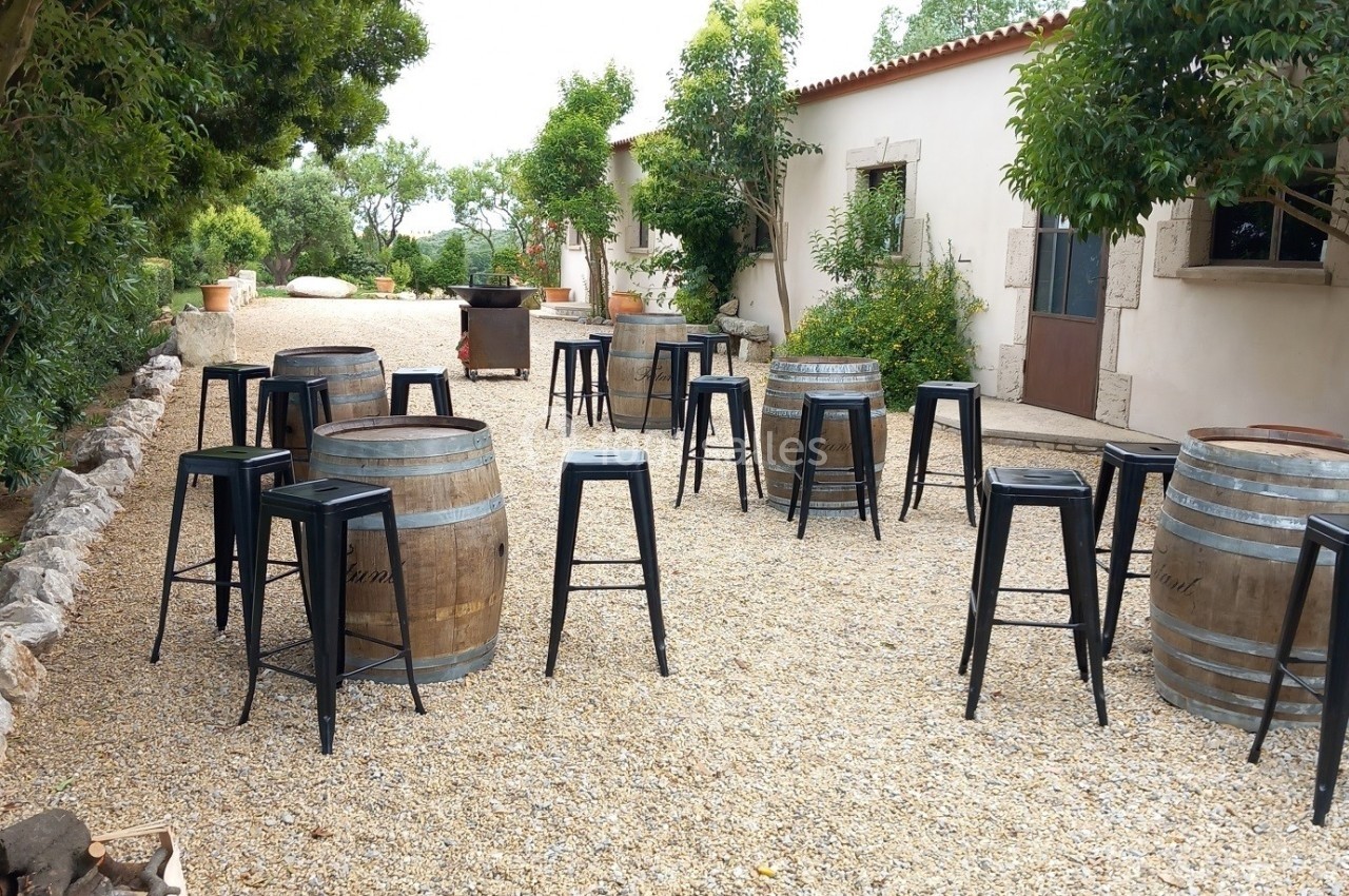 Cour extérieure avec des tonneaux en bois utilisés comme tables, entourés de tabourets noirs sur un sol en gravier.