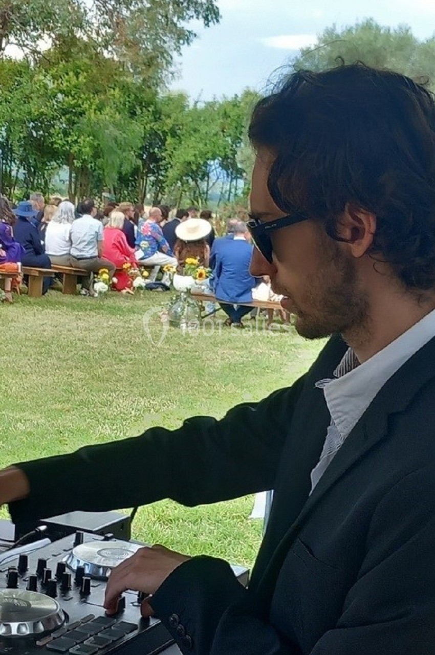 Un DJ mixant en plein air lors d'un événement avec des invités assis sur des bancs dans un cadre verdoyant.