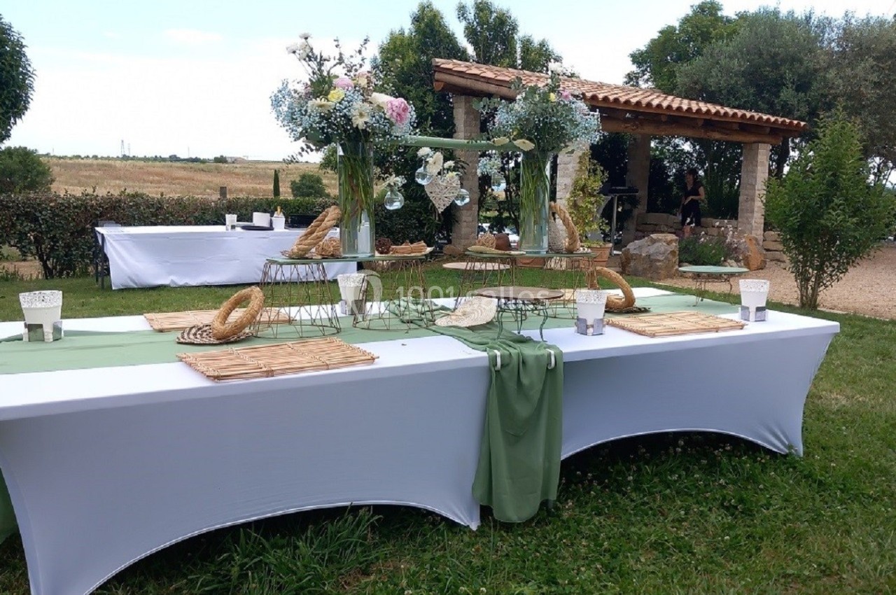 Tables dressées en extérieur avec nappes blanches et chemins de table verts, décorées de fleurs et paniers en osier.