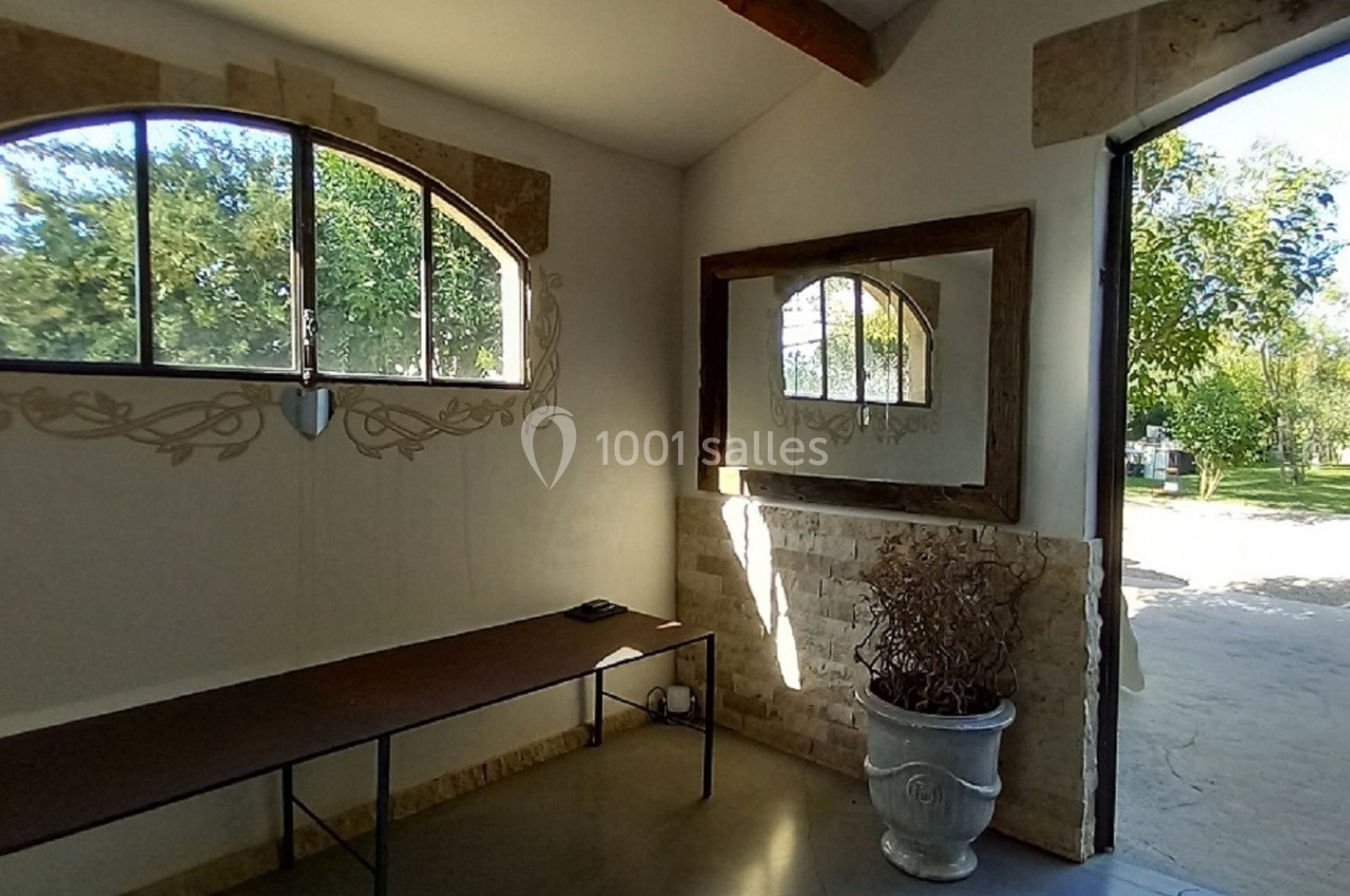 Intérieur lumineux avec table contre un mur, grande fenêtre et miroir, vue sur un jardin par une porte ouverte.
