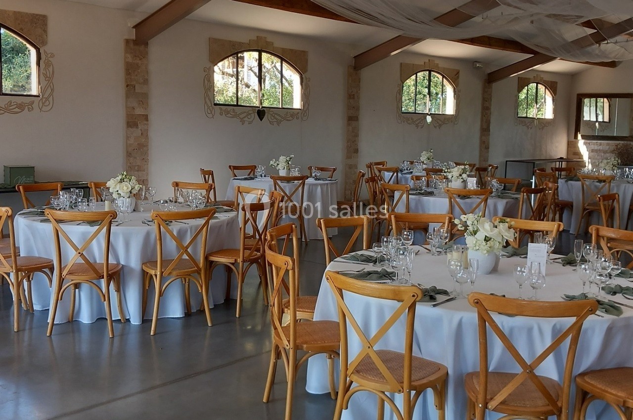 Salle de réception lumineuse avec tables rondes dressées, chaises en bois et décorations florales blanches.