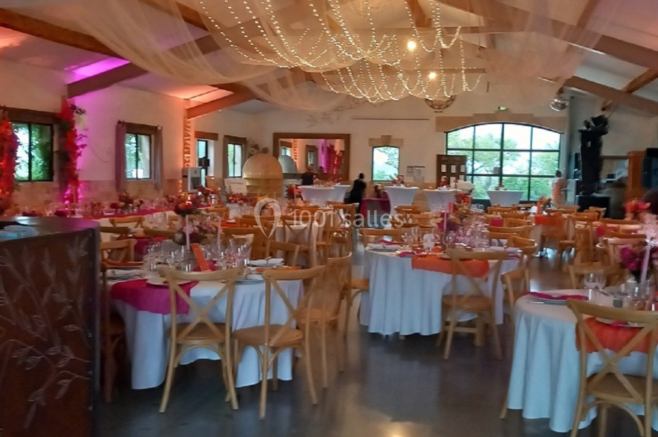 Salle de réception décorée avec des tables rondes, nappes blanches, chaises en bois et éclairage chaleureux.