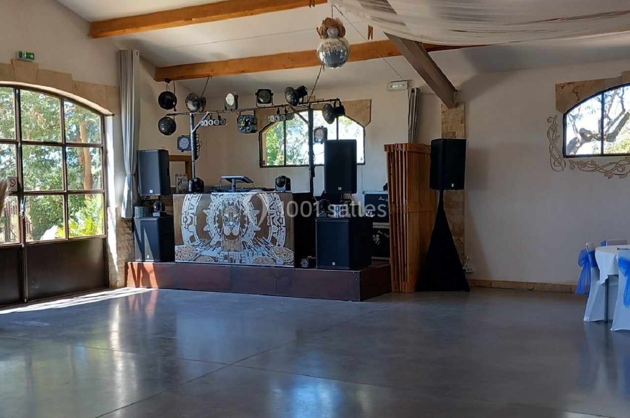 Salle de réception lumineuse avec piste de danse, tables décorées de rubans bleus et une scène équipée de matériel audio.