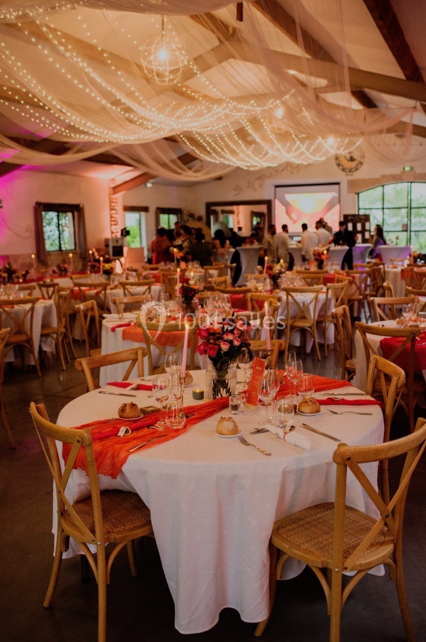 Salle de réception décorée avec des tables rondes, nappes blanches, chemins de table rouges et éclairage chaleureux.