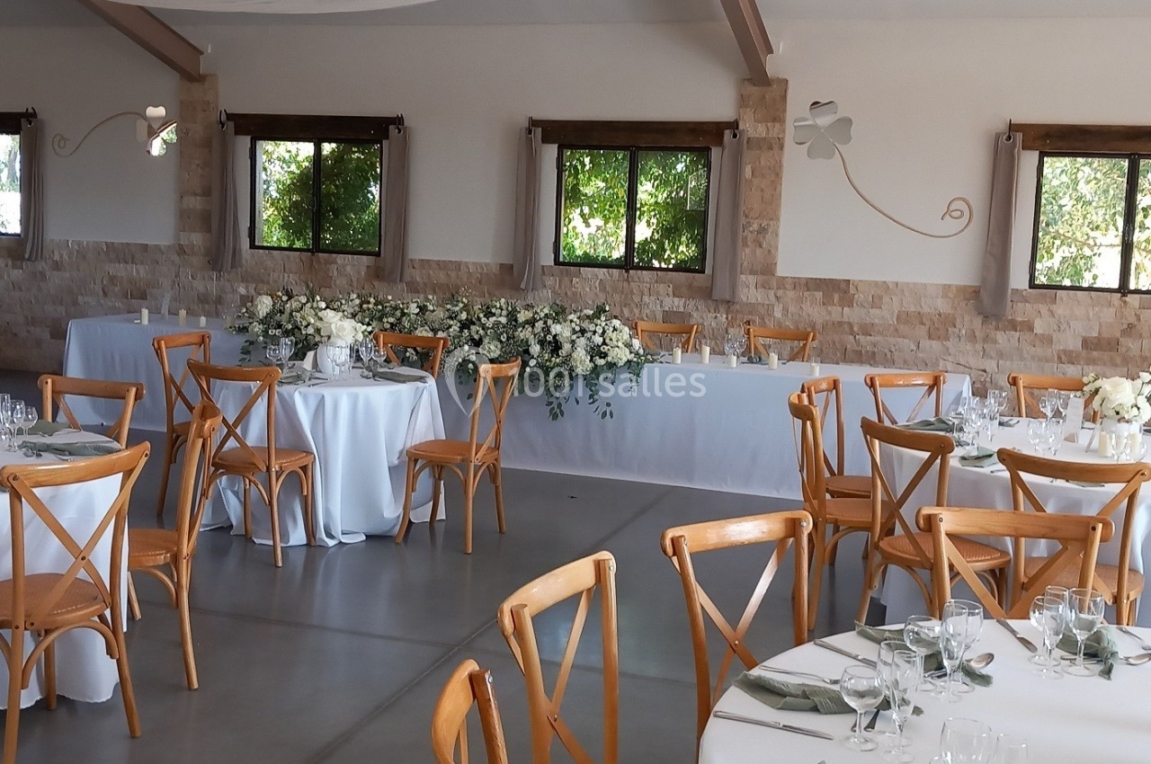 Salle de réception lumineuse avec tables rondes dressées, chaises en bois et décoration florale blanche et verte.