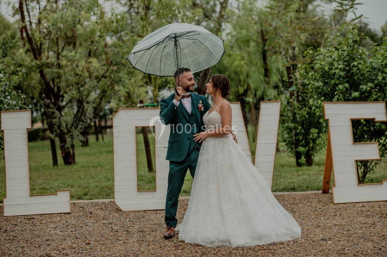 Un couple en tenue de mariage sous un parapluie transparent, devant des lettres formant le mot ’LOVE’ dans un jardin.