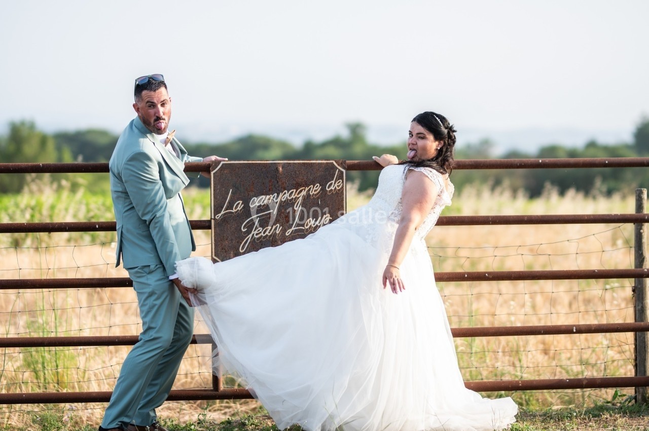 Un couple en tenue de mariage pose de manière décontractée devant une barrière métallique dans un champ.