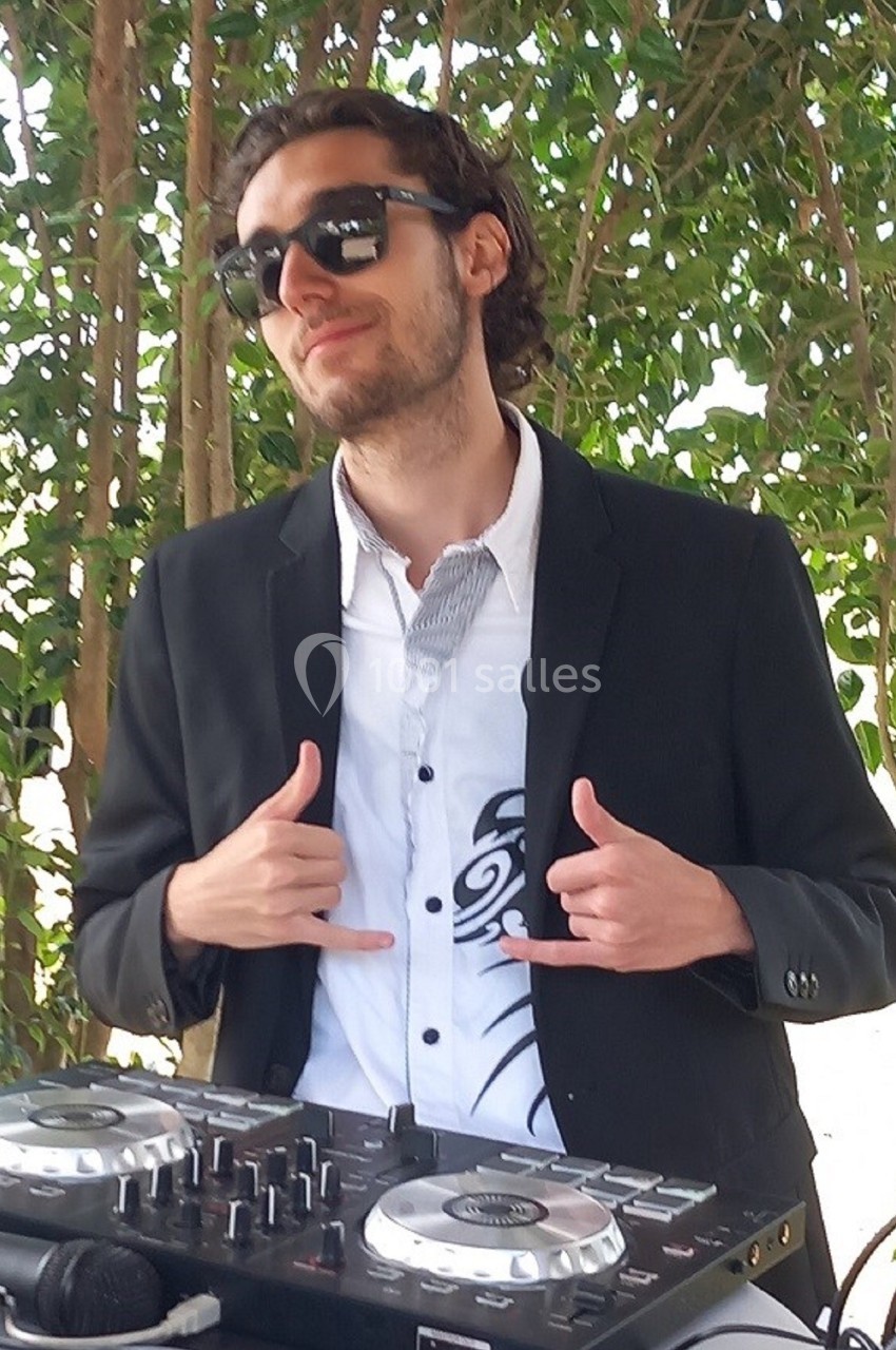 Un homme en costume noir et lunettes de soleil pose devant une table de mixage, avec des arbres en arrière-plan.